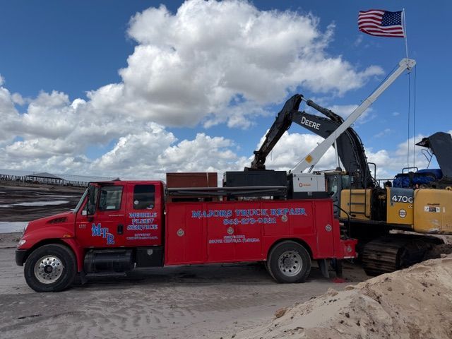 A red service truck parked at a sandy construction site next to a yellow Deere excavator with an American flag attached.