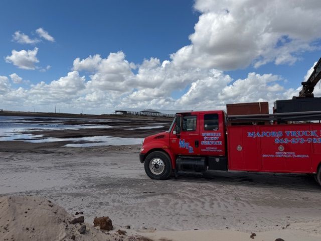 A red service truck parked on a sandy beach near a shoreline under a bright, cloudy blue sky.