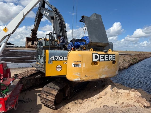 A John Deere 470G excavator sits on a dirt bank next to a canal, with its engine compartment open for maintenance.