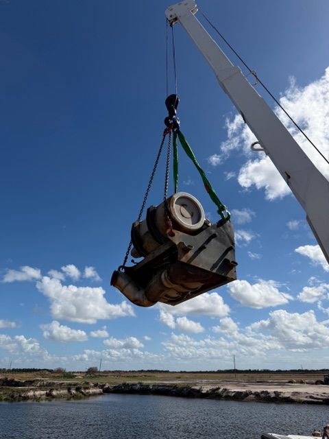 A crane arm suspends a heavy industrial metal assembly with cylindrical components above a body of water against a blue sky.