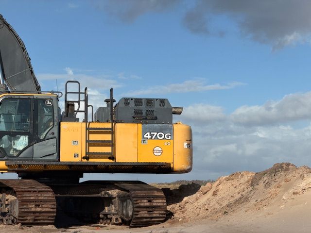 A yellow John Deere 470G excavator sits on a dirt site against a cloudy blue sky.