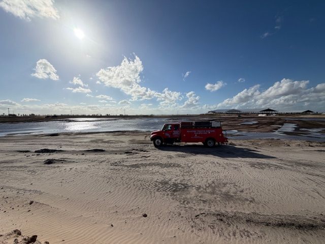 A red service truck parked on a vast, sandy, open landscape under a bright, sunny sky with scattered clouds.