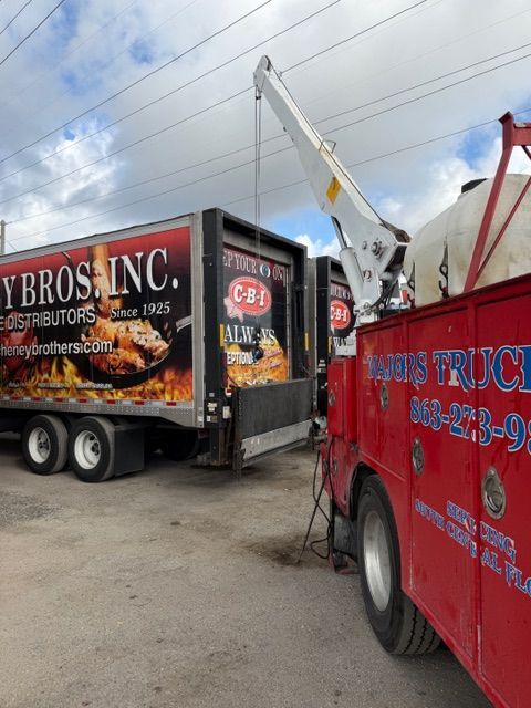 A white crane on a red truck bed positioned next to the back of a large commercial trailer marked 