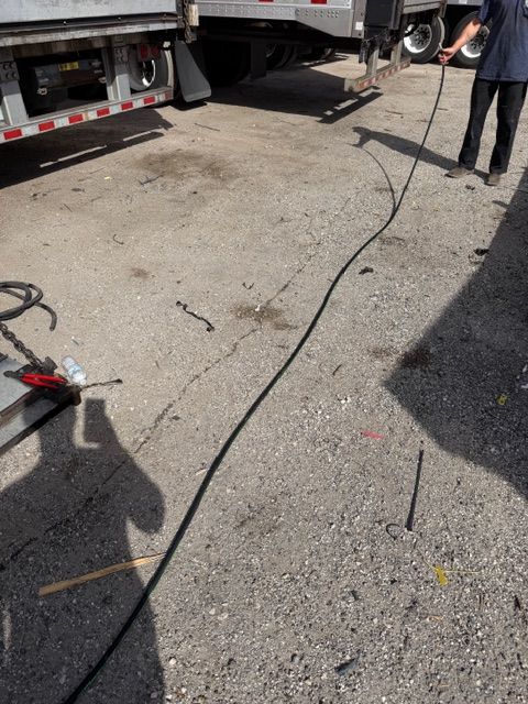 A person holds a long black cable extended across a gravel lot near a parked semi-trailer.