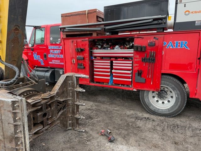 A bright red mobile repair truck with open tool cabinets and compartments parked on a dirt lot next to excavator parts.