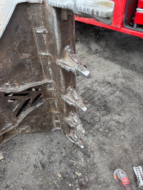 A metal excavator bucket rests on the ground, highlighting the teeth welded to its leading edge.