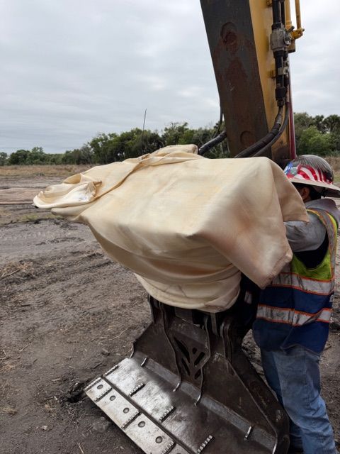 A worker in safety gear stands next to a large excavator attachment covered by a light-colored, draped fabric.