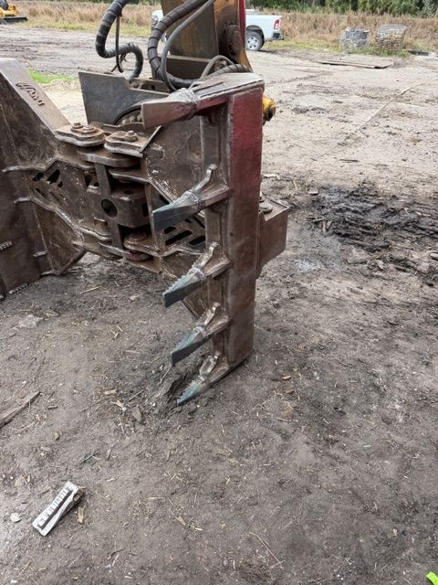 A heavy-duty metal grapple attachment for construction equipment rests on a dirt ground, showing four steel tines.