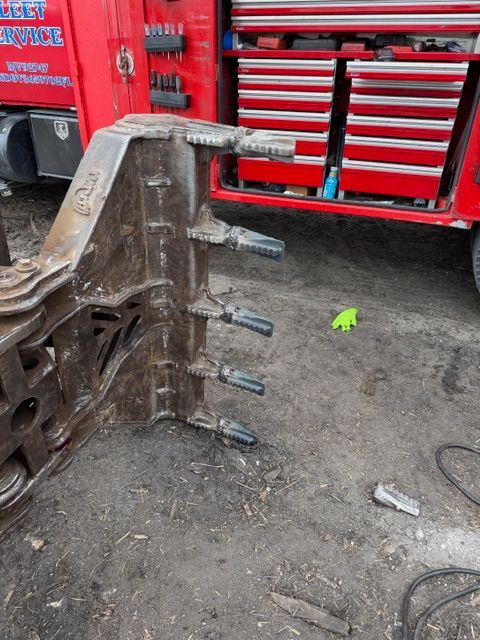 An excavator rake attachment sits on the dirt in front of an open, red mechanic’s truck tool chest.
