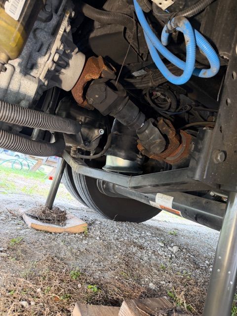 Underside of a vehicle showing a metal drive shaft, universal joints, and blue air lines near the rear wheels on gravel.