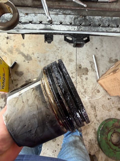 A person holds a heavily carbon-fouled engine piston with stuck and worn rings in a garage setting.