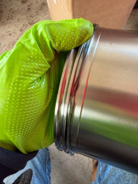 A gloved hand holds a cylindrical metal canister with a distinct ridge near the top.
