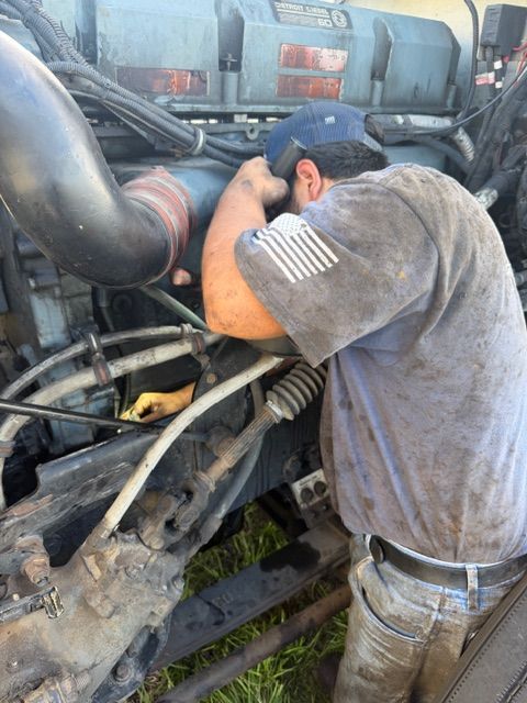 A mechanic in a gray shirt works on a large truck engine, leaning into the engine bay to inspect components.