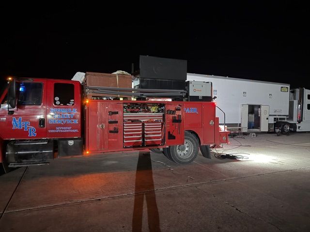 A red utility truck with open storage compartments and a large work trailer parked on a concrete lot at night.