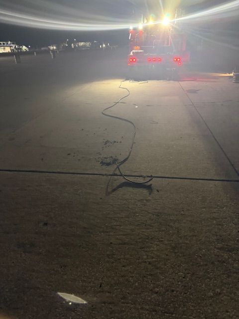 A truck with bright headlights and red taillights illuminates a dark paved area, with a long cable lying on the ground.