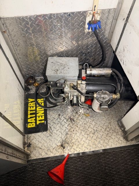 A hydraulic pump unit with hoses and a filter mounted on a diamond-plate floor near a Battery Tender and a red funnel.