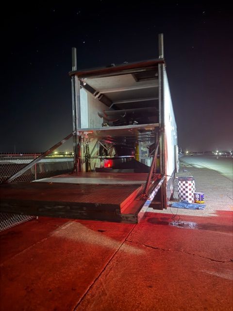 A race trailer at night with its rear ramp lowered, revealing a multi-level storage interior under floodlights.