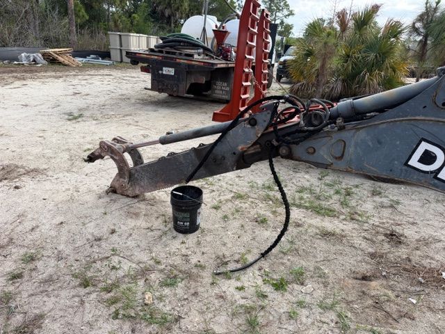 Rusty excavator arm with bucket in a sandy yard, with a work truck and red equipment in the background