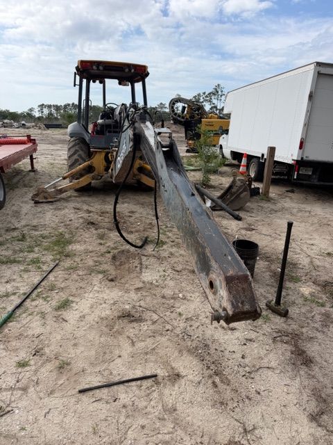 Backhoe lifting a long steel beam at a dirt worksite beside a white truck