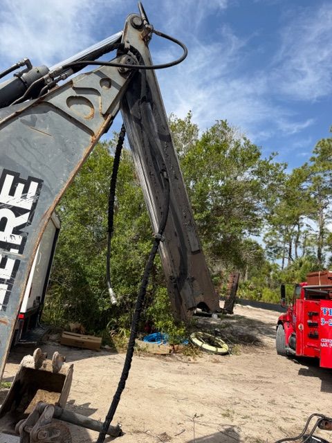 Excavator boom and hydraulic arm with dangling hose at a construction site, red machine in background