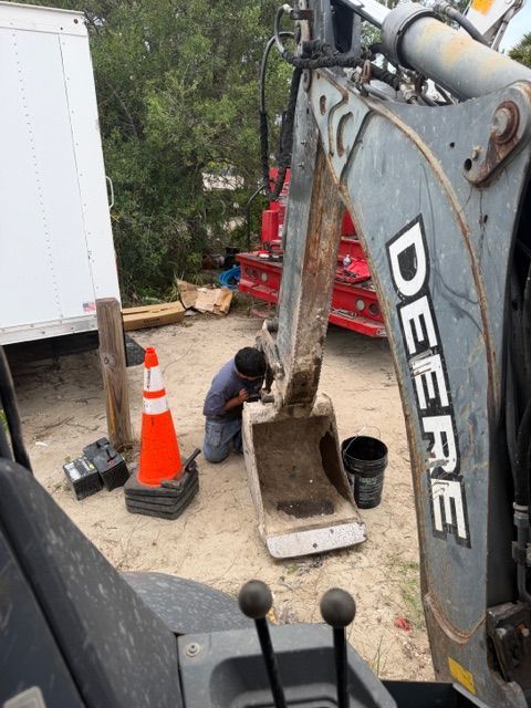 Excavator digging a trench beside a worker, orange cone, and truck in a yard.