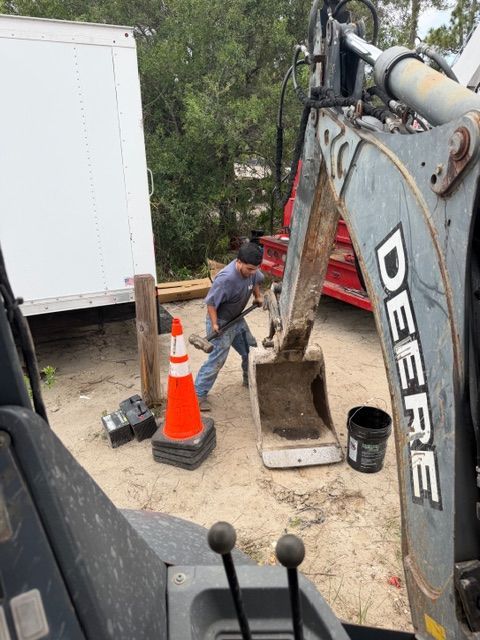 Worker in red shirt beside excavator near traffic cone and white truck on a dirt worksite
