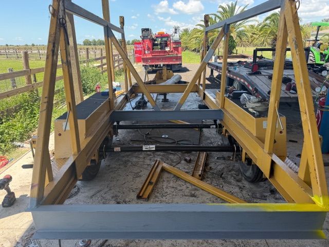 Yellow industrial metal frame on a trailer with a red machine in the background, outdoors on a road