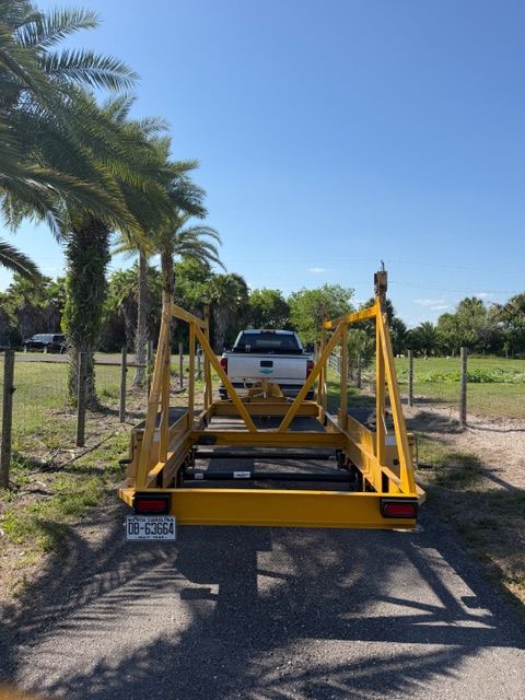 Yellow car trailer attached to a white pickup on a rural dirt road, with palm trees and fencing nearby
