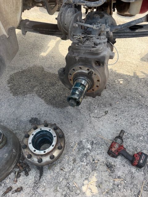 Disassembled vehicle axle and hub parts on concrete, with a red-and-black impact wrench nearby.