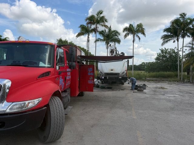 Red tow truck beside a white boat on a trailer in a palm-lined lot