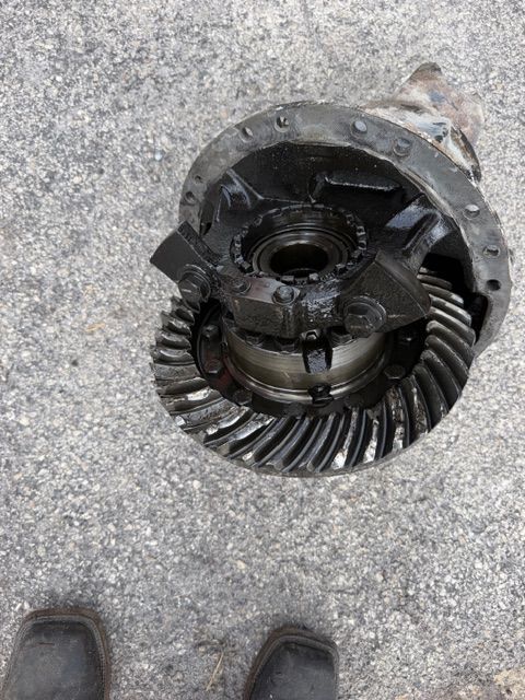 Broken metal differential gear lying on concrete pavement, with a shoe visible at the lower left.