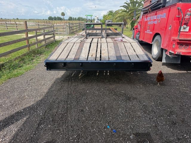 Flatbed trailer with wooden deck on a gravel road beside a red truck and fence in a rural area