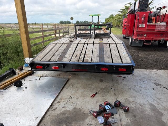 Flatbed trailer parked on a concrete pad beside a red truck, with wooden planks and a fenced field in the background
