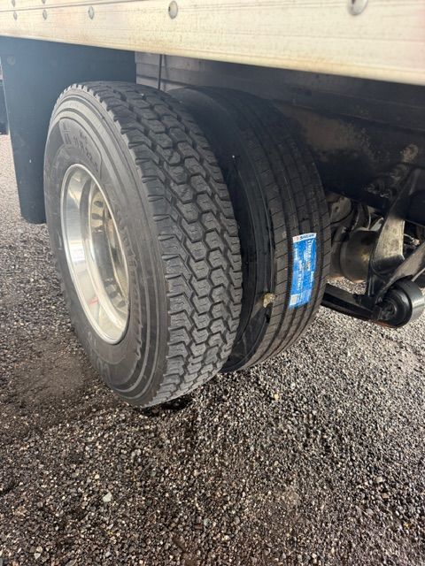 Dual truck tires under a trailer, with a blue label on the tread, parked on wet gravel.