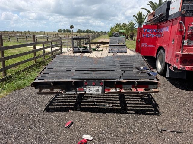 Flatbed truck trailer overturned on a rural road beside a red tow truck and wooden fence.