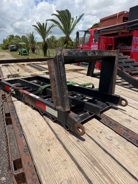 Black metal trailer hitch or tow bar on a wooden deck with palm trees and a red truck in the background