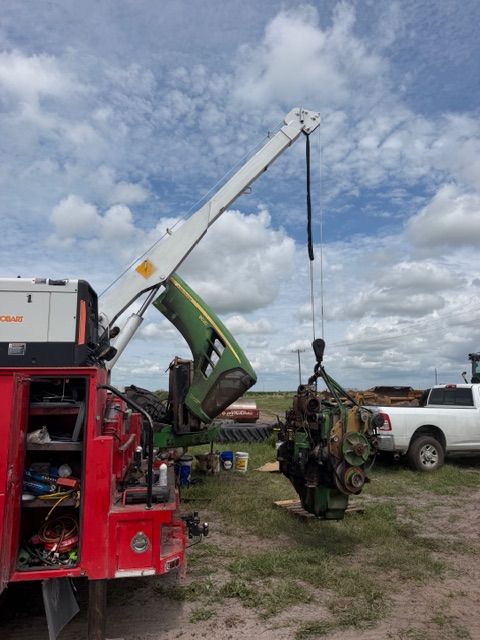 Red tow truck crane lifting a green tractor in a grassy yard beside a white pickup under cloudy sky