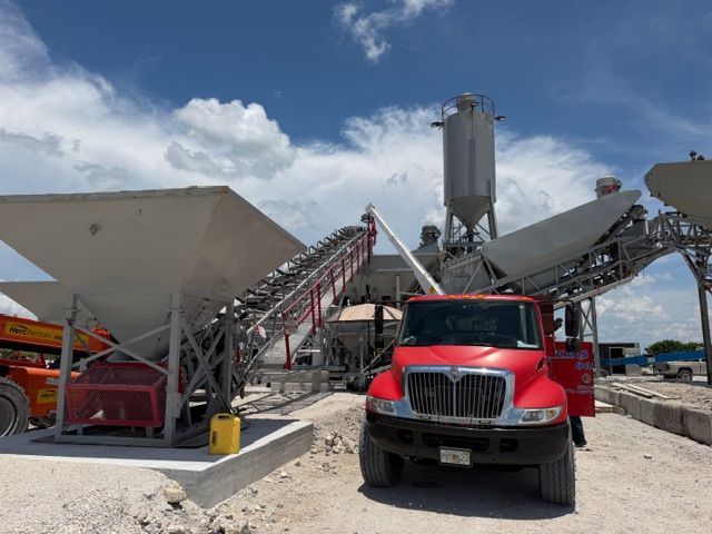 Red truck at a concrete plant with conveyors, silos, and hopper bins under a blue sky