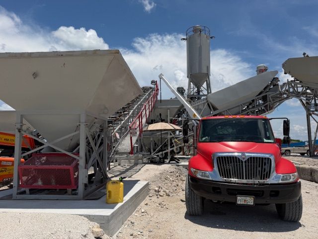 Red truck at a concrete plant with conveyors, silos, and white aggregate bins under a blue sky