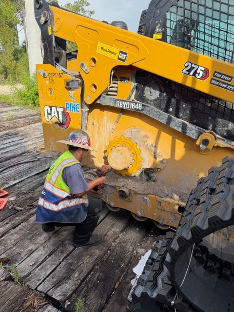Worker in hard hat kneels beside a yellow CAT machine on a muddy construction site