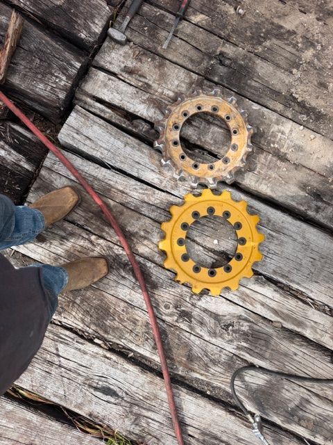 Two yellow gear-like metal parts on weathered wooden planks next to a red hose and a person’s boots