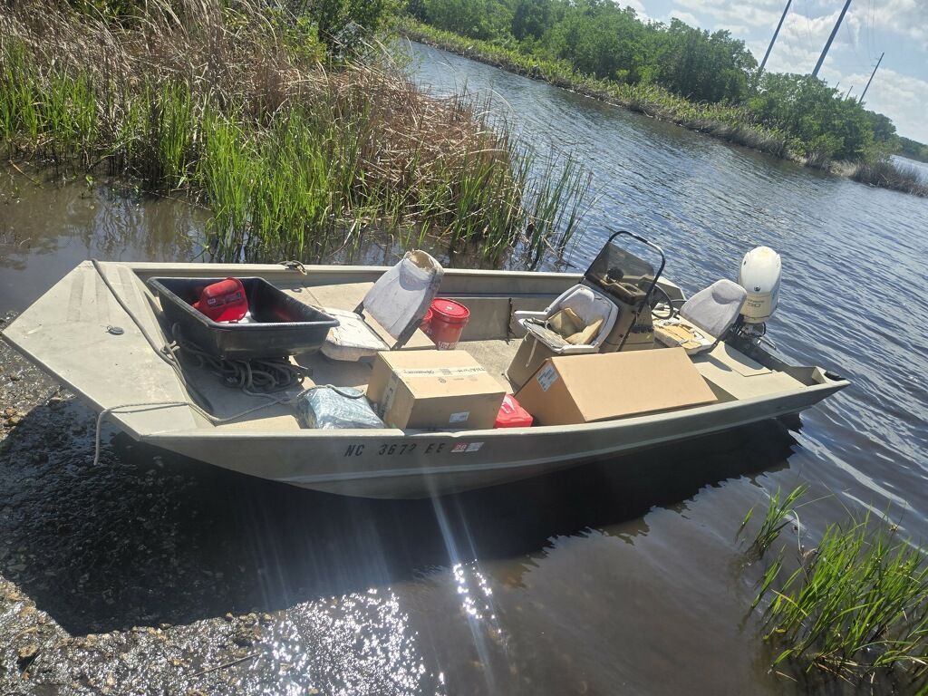 Small aluminum boat with fishing gear on a calm river beside grassy banks and reeds
