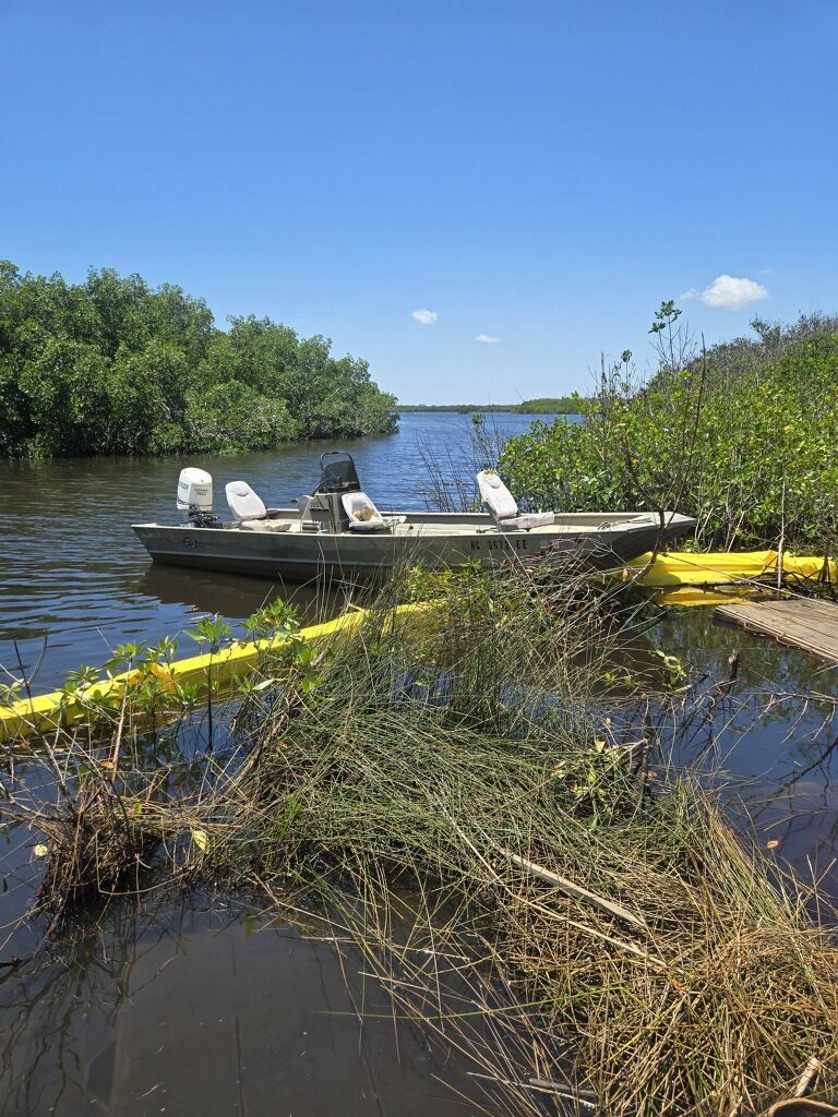 Small motorboat in a marshy river, surrounded by yellow floating booms and green trees under a blue sky.