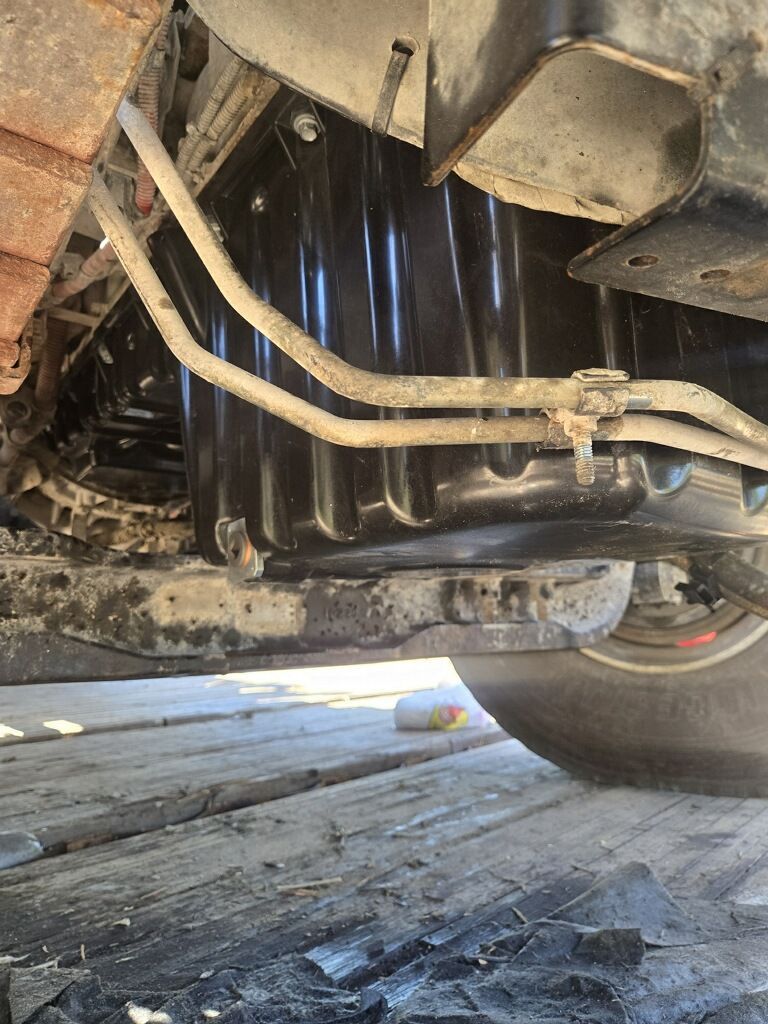 Underbody view of a vehicle showing black oil pan and metal lines over a wooden floor