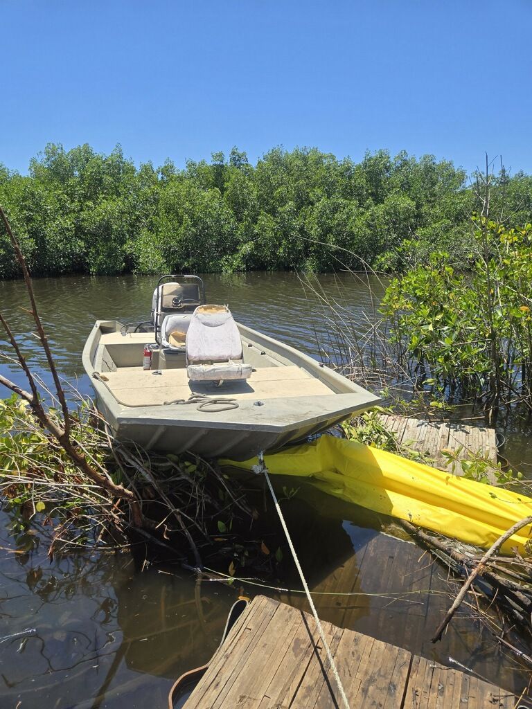 Small boat grounded by a muddy riverbank, next to a yellow kayak and dock planks, under a clear blue sky