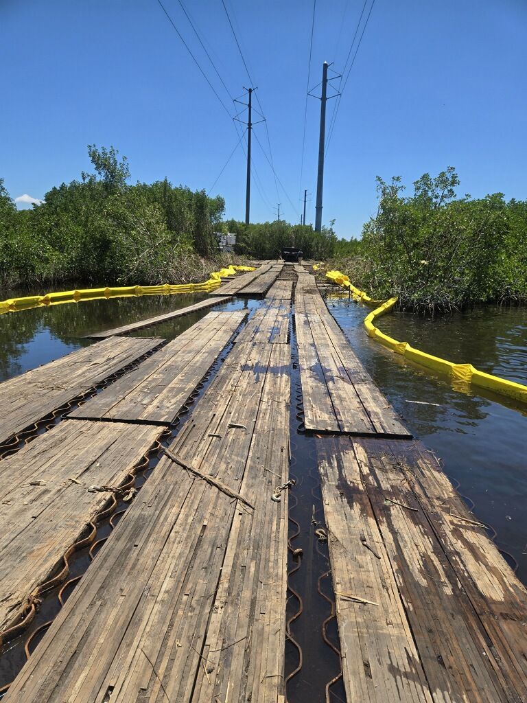 Weathered wooden boardwalk through shallow water, bordered by yellow floating booms under a clear blue sky.