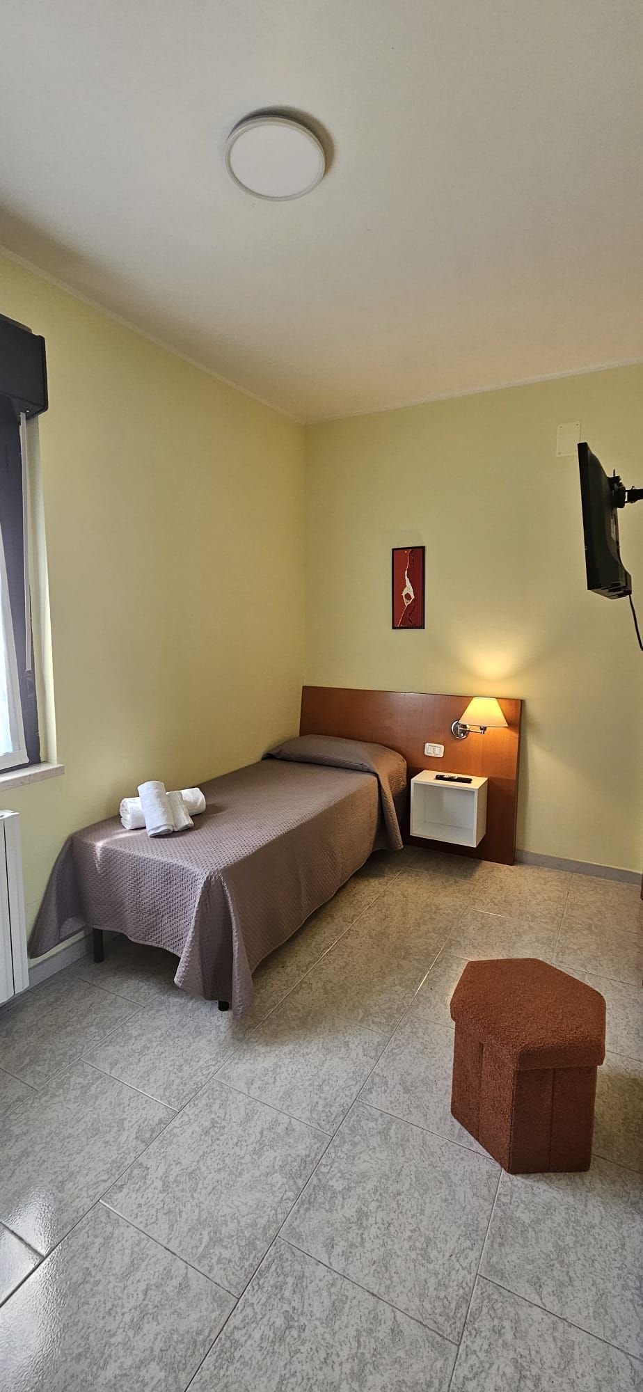 Piccola camera d'albergo con un letto singolo, un comodino e un piccolo sgabello. Le pareti sono gialle e il pavimento è piastrellato.