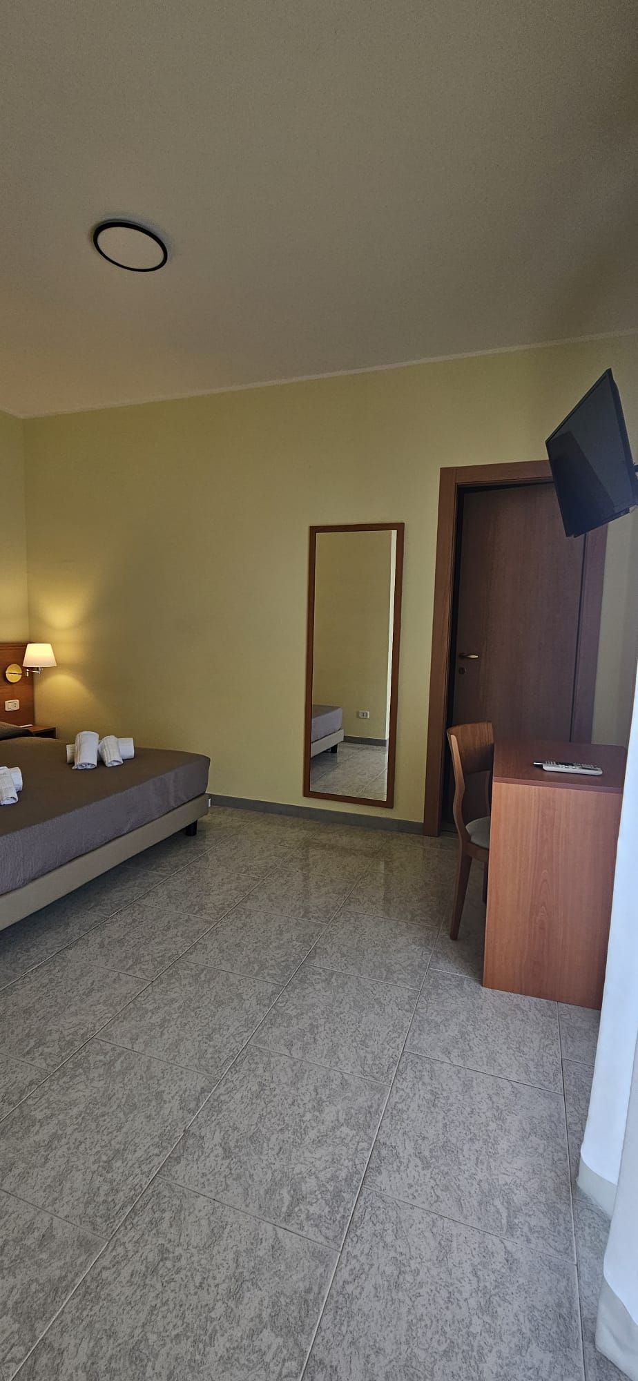 Camera d'albergo con letto, specchio, scrivania e TV. Pareti gialle, pavimento a motivi e una porta marrone scuro.
