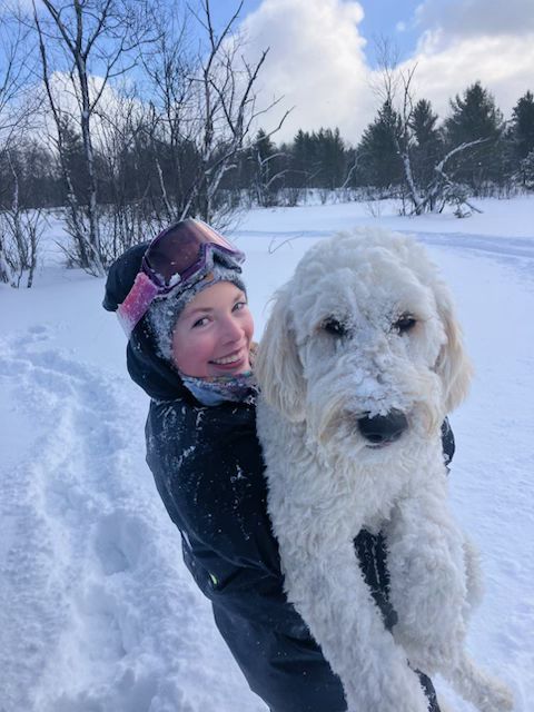 A woman wearing goggles is holding a white dog in the snow.