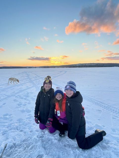 Three people are posing for a picture in the snow with a dog in the background.